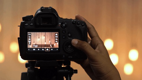 Photography of a close-up of a person adjusting settings on a DSLR camera mounted on a tripod, with blurred warm lights in the background.