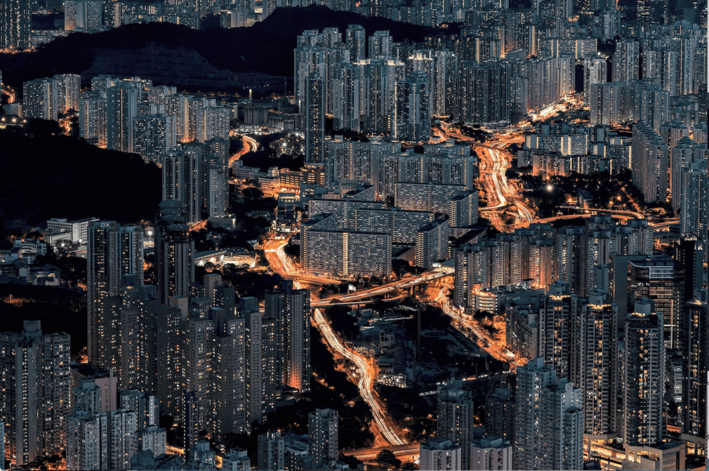 “A dense modern cityscape at night with high-rise apartment buildings and illuminated roads forming flowing light trails through the urban area.”