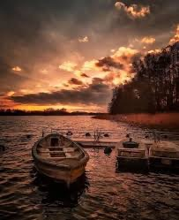 “Old wooden boats floating on a calm lake at sunset, with dramatic orange clouds reflecting on rippling water and a dark tree-lined shore in the background.”