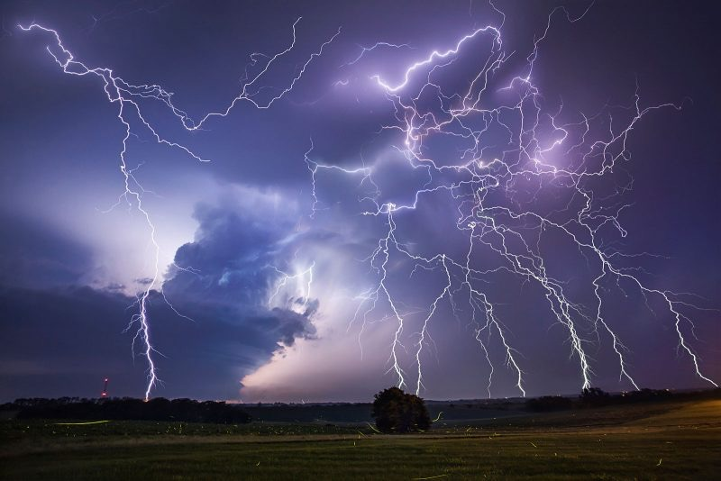 “A wide open field under a stormy sky filled with multiple branching lightning strikes illuminating dark clouds and the horizon at dusk.”