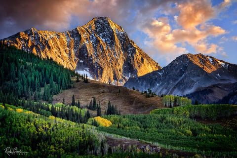 “Sunlit rocky mountain peaks rising above dense green forest, with golden light hitting the cliffs under a partly cloudy sky.”